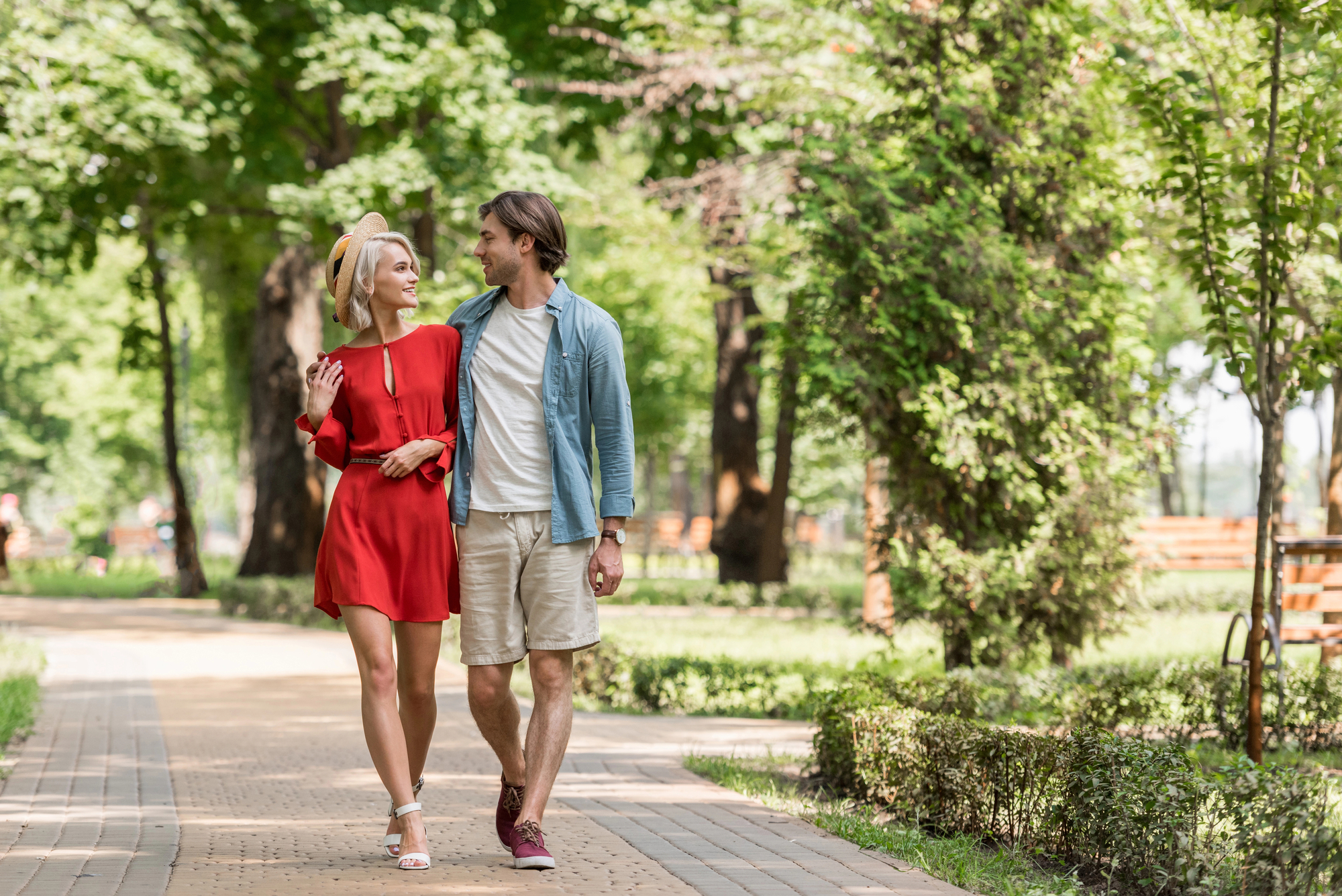 A man and woman smile at each other as they walk arm in arm down a tree-lined path in a park. The woman wears a red dress and sandals, and the man wears a light blue shirt over a white t-shirt with beige shorts and red sneakers. The park is lush and green.