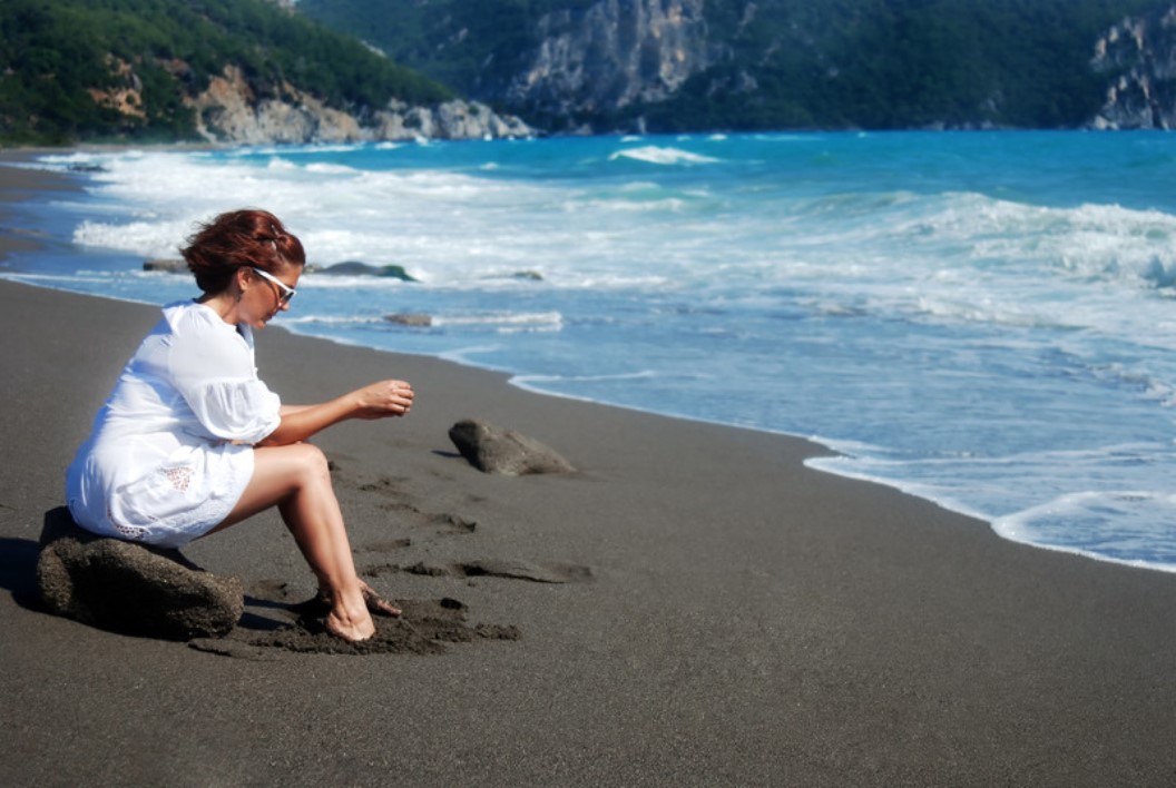 A person wearing a white shirt sits on a rock at the edge of a sandy beach, facing the ocean. They are holding something in their hands and looking towards the water. The sea is a vibrant blue, and lush green hills can be seen in the background.