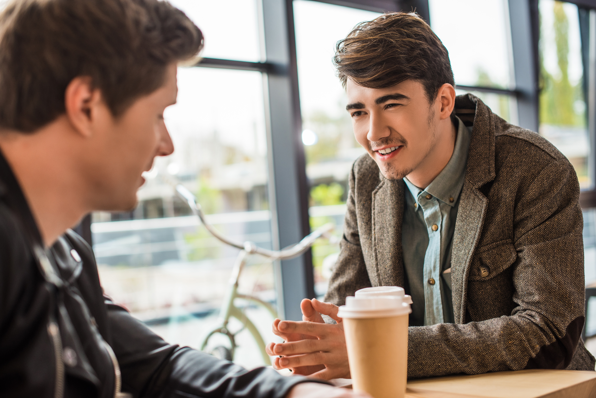 Two young men are sitting at a table near a window, engaged in a conversation. One man with short brown hair smiles while talking, wearing a blazer over a button-up shirt. The other man, facing away from the camera, wears a dark jacket. A take-out coffee cup sits on the table.