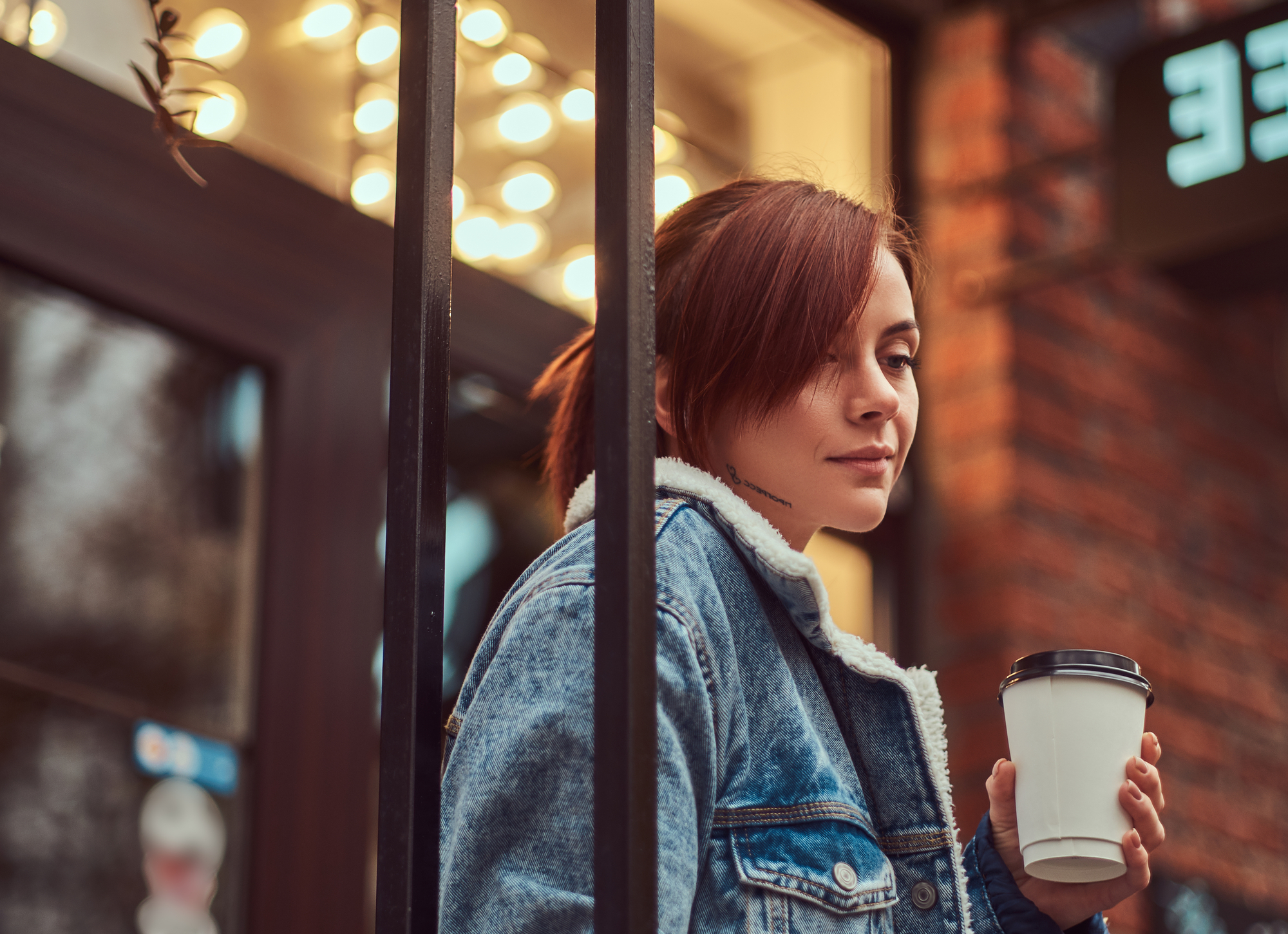 A person with short brown hair, wearing a denim jacket, holds a white to-go coffee cup while standing outside near a metal railing. Warm lights and a brick wall in the background create a cozy atmosphere.