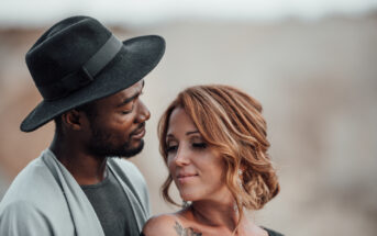 A woman and man stand close together in an intimate pose. The man, wearing a dark hat and gray jacket, gazes lovingly at the woman. The woman, with red hair and a tattoo on her shoulder, looks down with a soft smile. The background is blurred and neutral-toned.