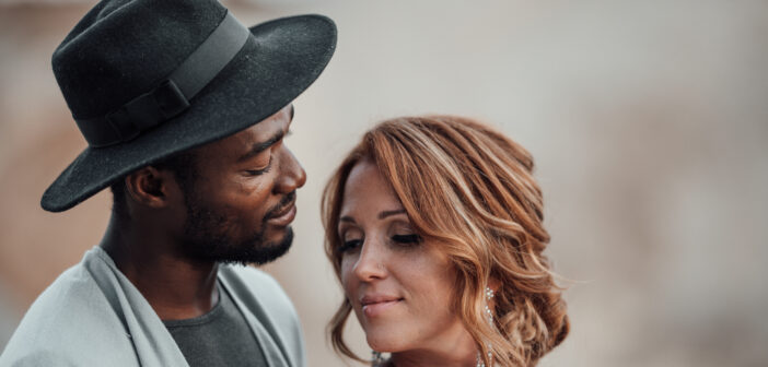 A woman and man stand close together in an intimate pose. The man, wearing a dark hat and gray jacket, gazes lovingly at the woman. The woman, with red hair and a tattoo on her shoulder, looks down with a soft smile. The background is blurred and neutral-toned.