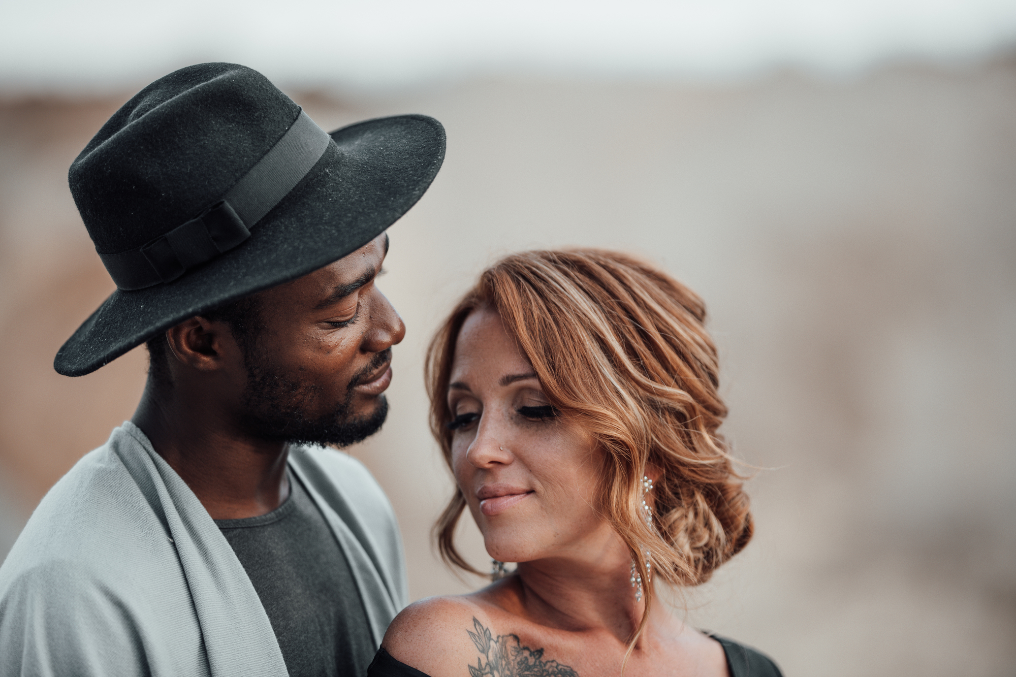 A woman and man stand close together in an intimate pose. The man, wearing a dark hat and gray jacket, gazes lovingly at the woman. The woman, with red hair and a tattoo on her shoulder, looks down with a soft smile. The background is blurred and neutral-toned.