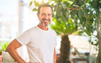 A middle-aged man with short hair and a beard smiles warmly while standing in a sunlit room filled with green plants. He is wearing a light-colored t-shirt and has one hand on his hip. Sunlight filters through the leaves, creating a bright, cheerful atmosphere.