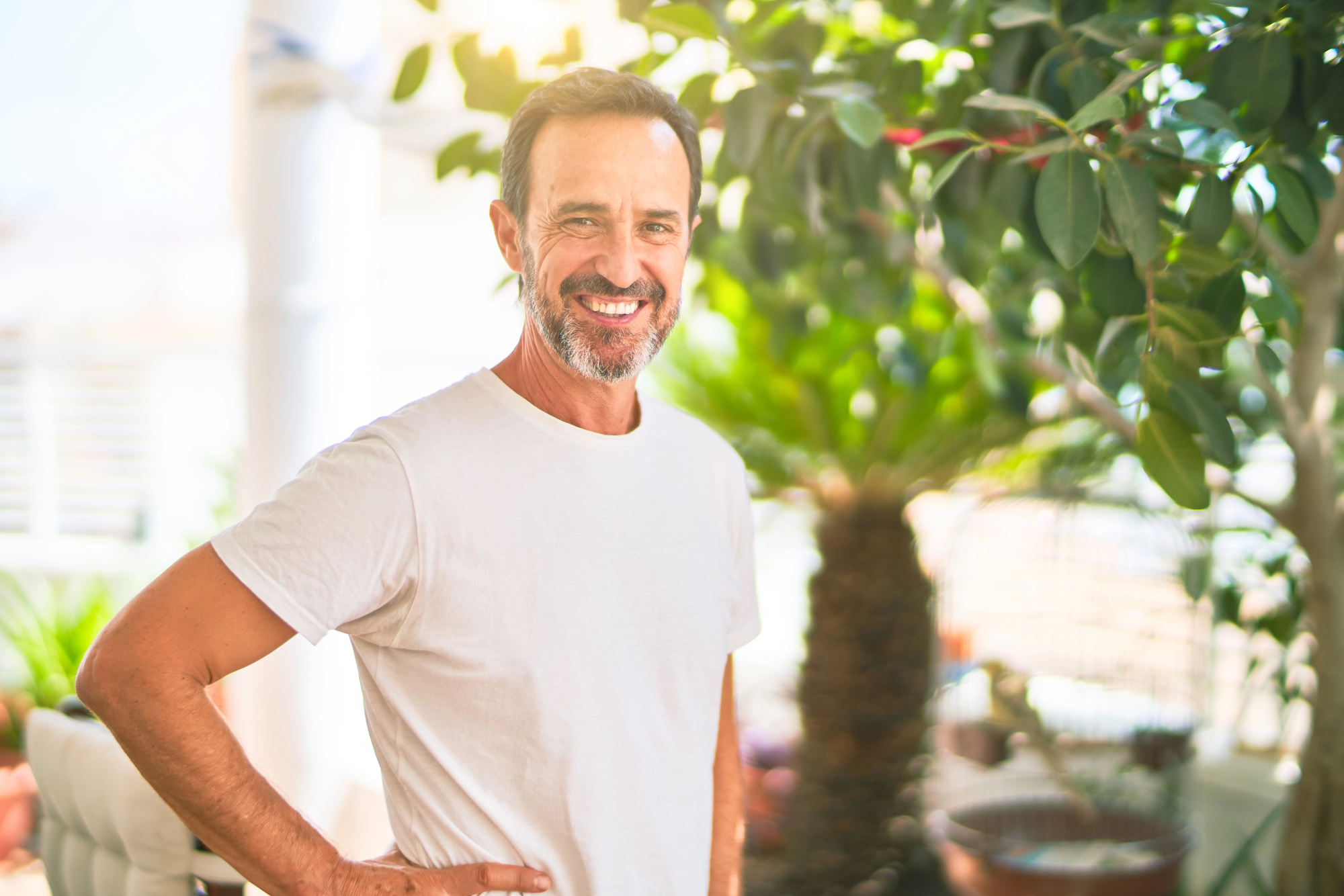 A middle-aged man with short hair and a beard smiles warmly while standing in a sunlit room filled with green plants. He is wearing a light-colored t-shirt and has one hand on his hip. Sunlight filters through the leaves, creating a bright, cheerful atmosphere.