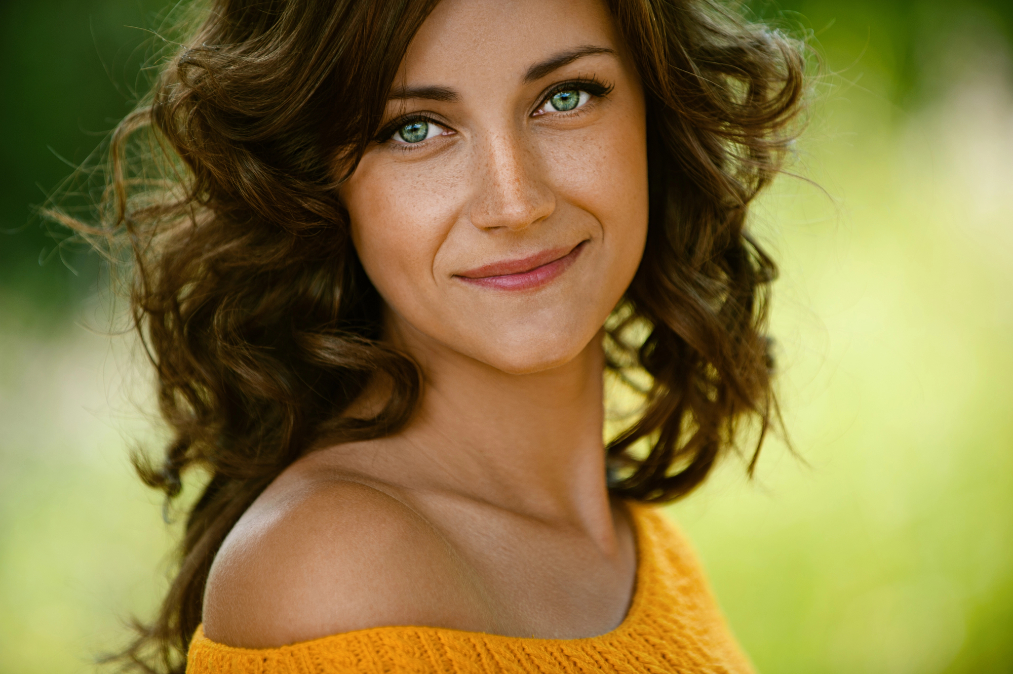 A woman with wavy brown hair and green eyes smiles warmly at the camera. She is wearing an off-shoulder yellow top. The background is a blurred natural setting with greenery, suggesting she is outdoors.