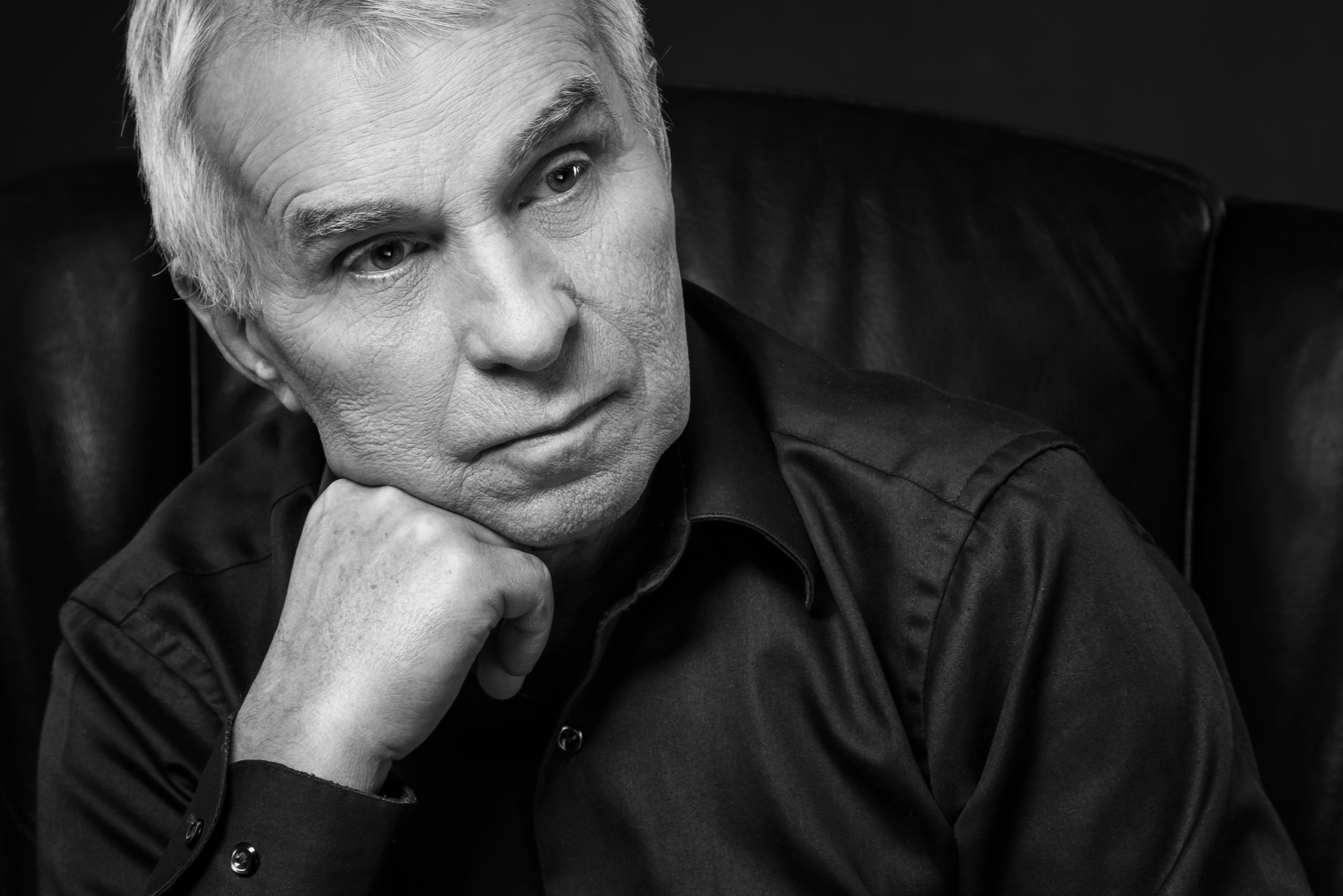 A grayscale image of an older man with light hair, sitting in a leather chair. He is wearing a long-sleeve button-up shirt and has a contemplative expression, with his chin resting on his hand and looking slightly to the side. The background is dark and plain.