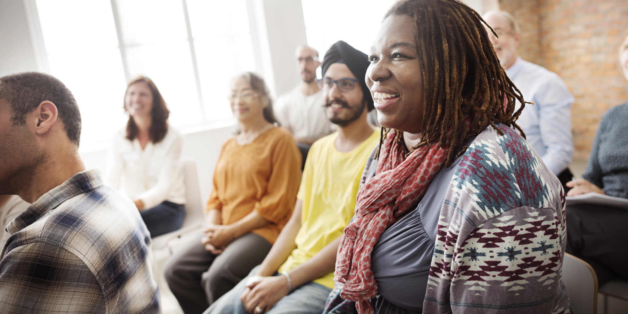 A diverse group of people sitting in a well-lit room, smiling and attentively listening to someone off-camera. The group appears engaged and cheerful, suggesting they are participating in a positive and interactive event or discussion.