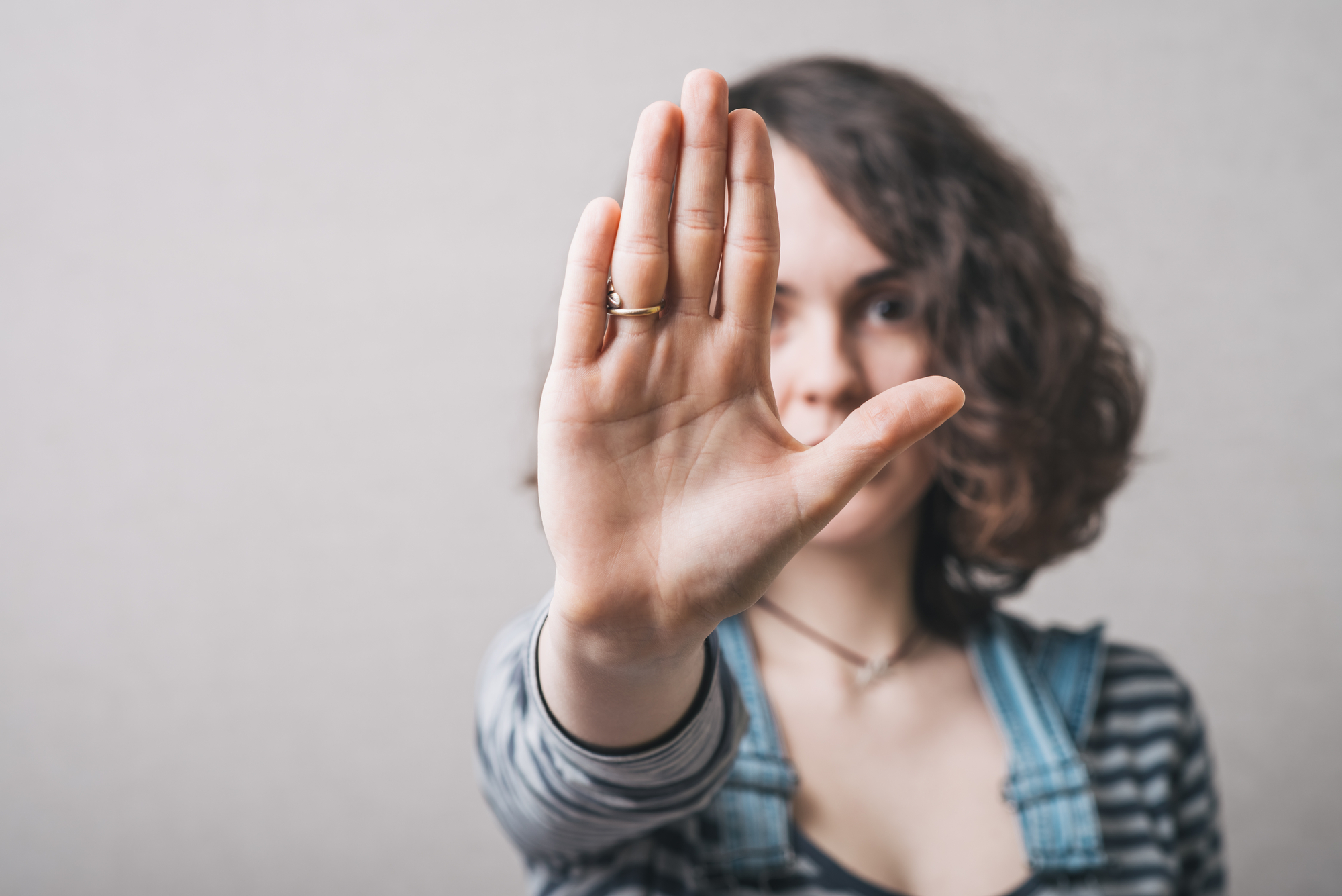 A person with curly hair is holding up their hand in front of the camera, creating a stop or no gesture. Their face is partially obscured by their hand. They are wearing a striped shirt and denim overalls against a neutral background.