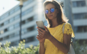 A woman wearing reflective sunglasses and a yellow lace dress stands outdoors while looking at her smartphone. She is backlit by sunlight, with a building and greenery in the background.