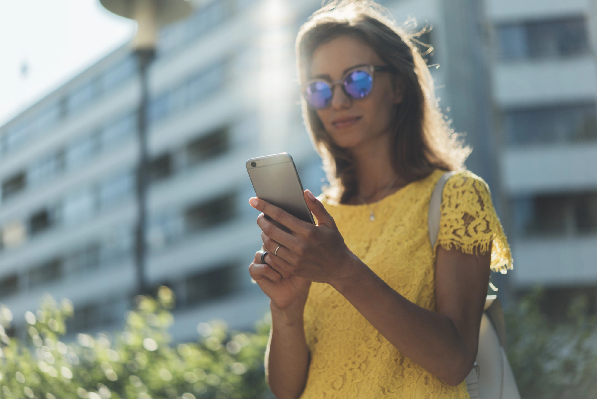 A woman wearing reflective sunglasses and a yellow lace dress stands outdoors while looking at her smartphone. She is backlit by sunlight, with a building and greenery in the background.