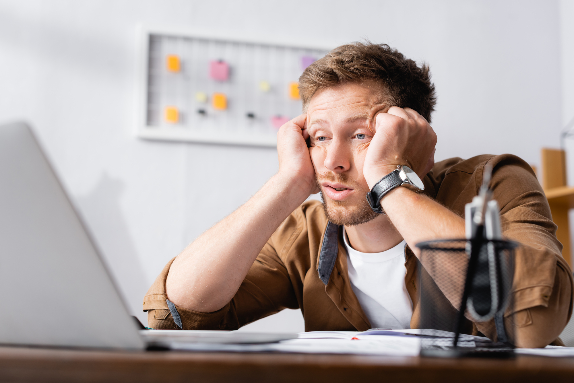 A man with a frustrated expression sits at a desk, staring at a laptop screen. He rests his head in his hands, with elbows on the desk. In the background, there's a board with sticky notes attached. A pen holder and some papers are on the desk in front of him.