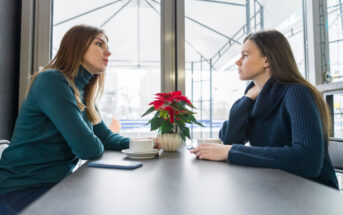 Two women sit across from each other at a table in a cafe, engaged in a serious conversation. Both are wearing sweaters. A potted poinsettia sits on the table between them, alongside two white coffee cups and a smartphone. Large windows are in the background.