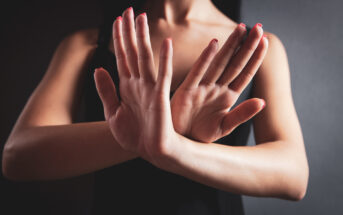 A person stands against a dark background with their hands crossed in front, palms facing outward, in a gesture of refusal or defense. They are wearing a sleeveless black top, and their fingernails are painted red. Their face is not visible.