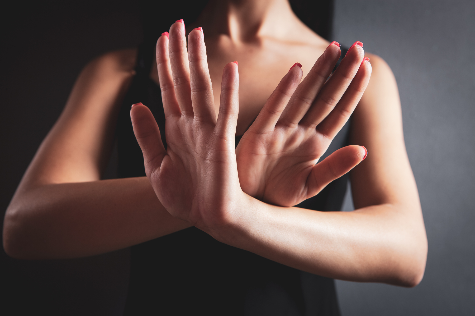 A person stands against a dark background with their hands crossed in front, palms facing outward, in a gesture of refusal or defense. They are wearing a sleeveless black top, and their fingernails are painted red. Their face is not visible.