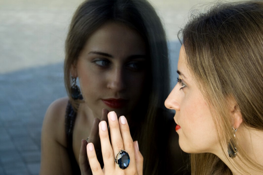 A woman with long brown hair gazes at her reflection in a glass surface, her hand touching the glass. She wears a large, dark gemstone ring and matching earrings, and has red lipstick. The reflection shows a similar pose, creating a contemplative mood.