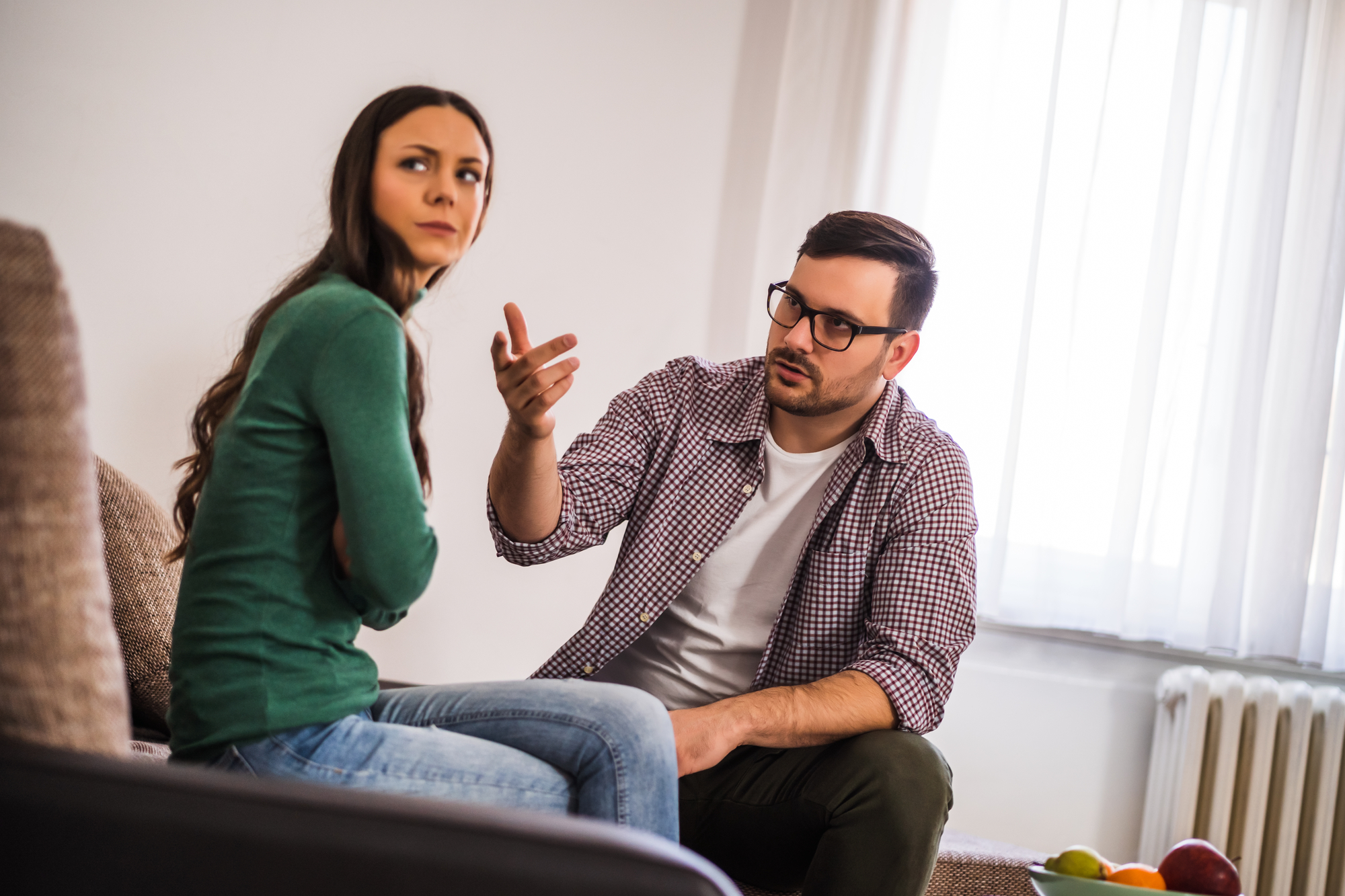 A man with glasses sits on a couch gesturing with his hand towards a woman with long brown hair, who looks away with her arms crossed. They appear to be having a serious conversation in a well-lit room.