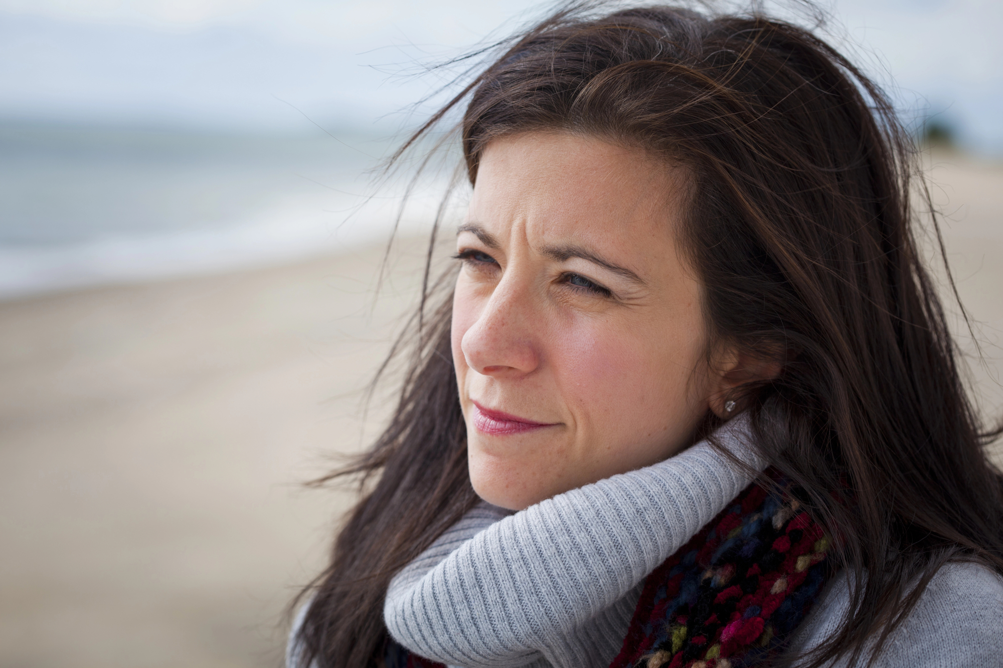 A woman with long brown hair stands on a beach, looking into the distance with a thoughtful expression. She is wrapped in a thick, gray sweater and a colorful scarf. The beach and ocean are blurred in the background under an overcast sky.