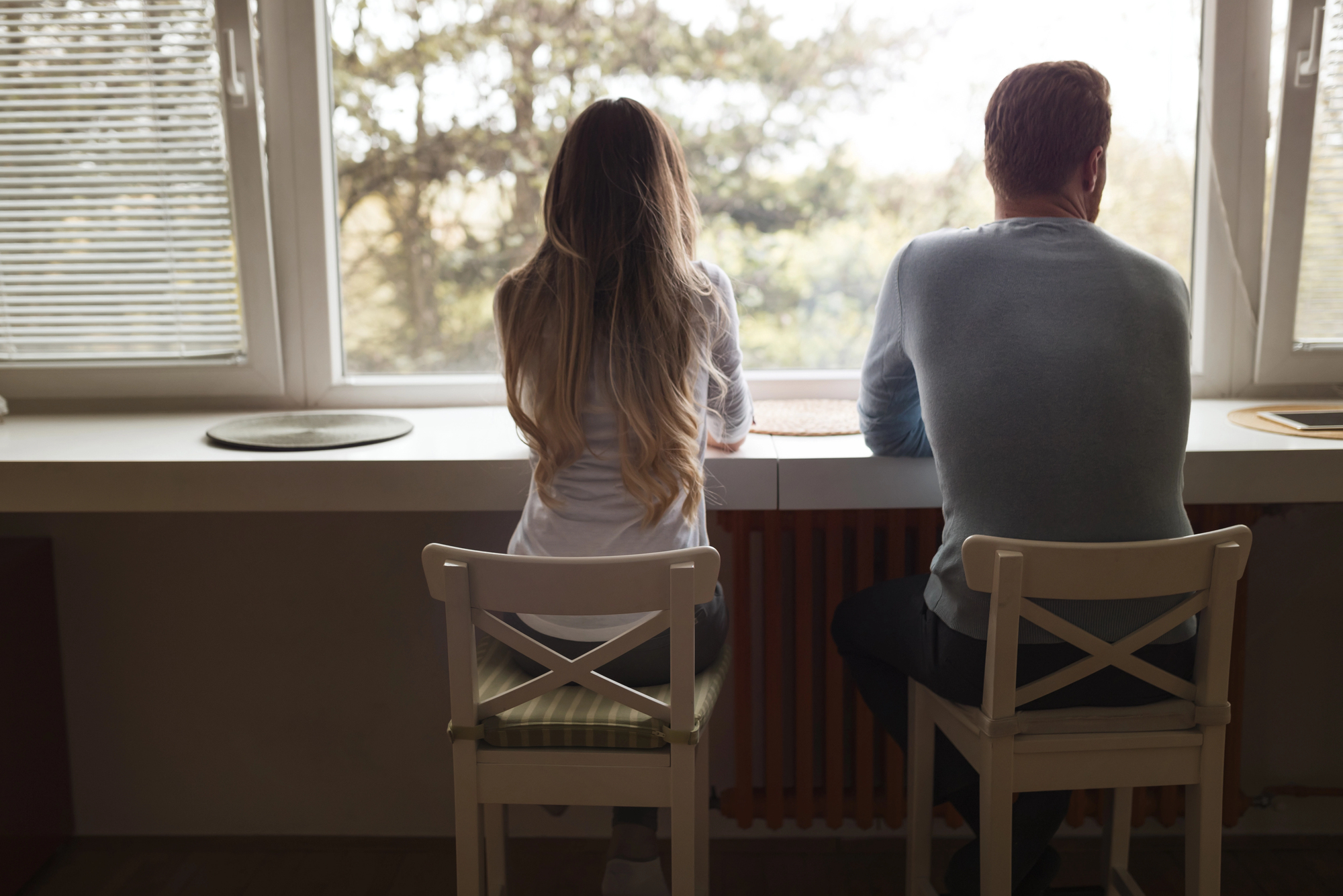 A woman and a man sit on white chairs, facing away from the camera and looking out of a large window. Sunlight streams in, illuminating their backs. Tall trees are visible through the window. Both wear casual clothing. The scene has a tranquil, contemplative atmosphere.