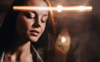 A woman with long, brown hair looks down with a serene expression. There's a warm light in the background and a light trail effect across the image, creating a calm and reflective atmosphere. She wears a plaid shirt and a delicate pendant necklace.