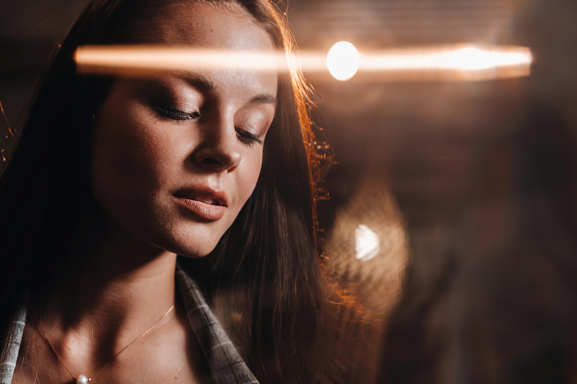 A woman with long, brown hair looks down with a serene expression. There's a warm light in the background and a light trail effect across the image, creating a calm and reflective atmosphere. She wears a plaid shirt and a delicate pendant necklace.