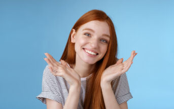 A smiling young woman with long red hair is wearing a striped t-shirt and holding her hands up near her face in a playful gesture. She is standing against a solid light blue background.