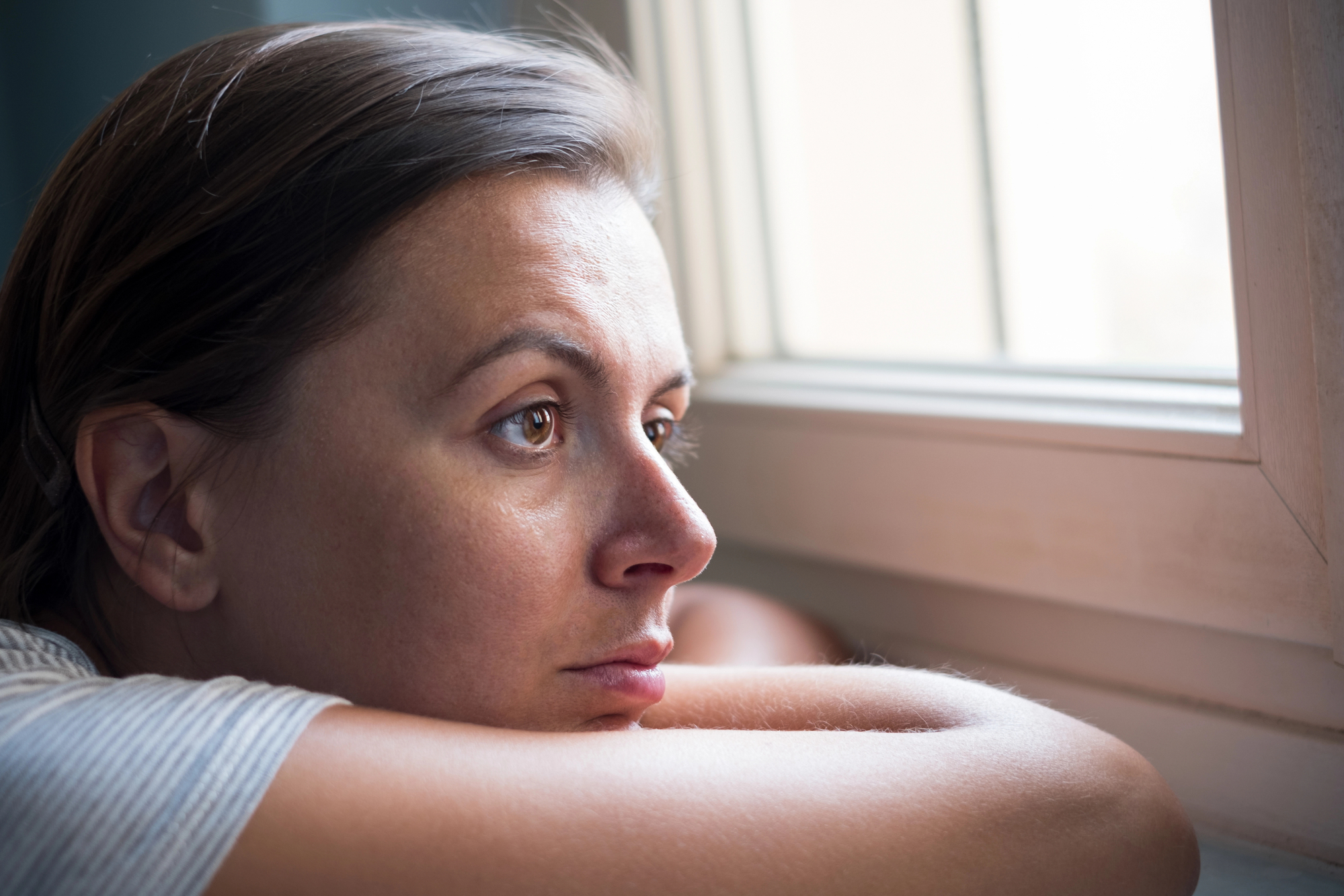 A woman with a thoughtful and slightly melancholic expression gazes out of a window. She rests her arms on the windowsill, leaning her head on them. Sunlight illuminates her face, creating a soft, introspective mood.