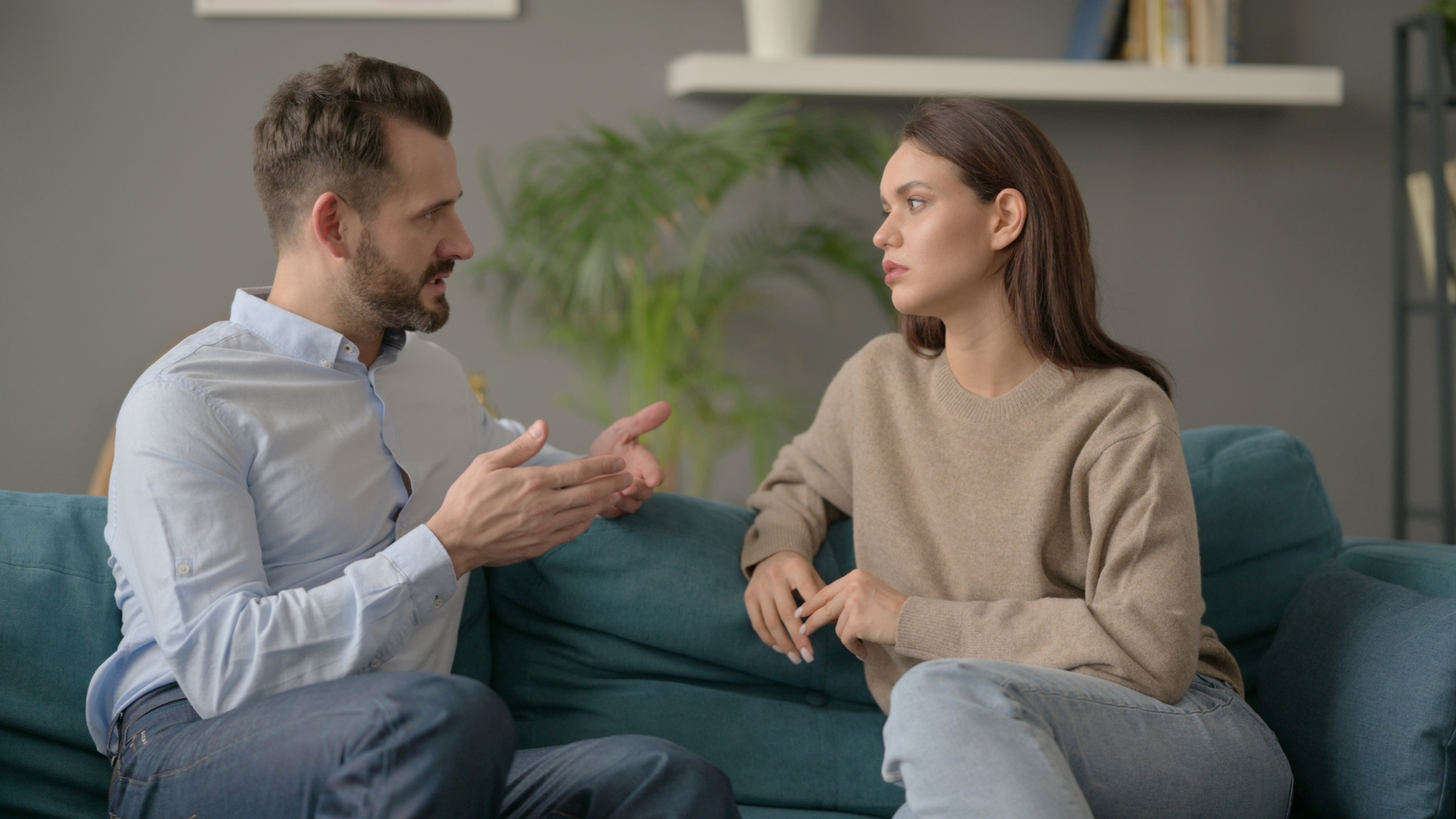 A man and a woman sit on a blue couch having an animated conversation. The man, gesturing with his hands, wears a light blue shirt and jeans, while the woman, listening intently, wears a beige sweater and jeans. A plant and a shelf with books are in the background.