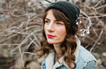 A person with long brown hair, red lipstick, and a black beanie stands outdoors in front of bare branches. They are wearing a denim jacket with a fleece collar and have a thoughtful expression. The branches have small buds on them.