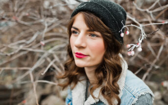 A person with long brown hair, red lipstick, and a black beanie stands outdoors in front of bare branches. They are wearing a denim jacket with a fleece collar and have a thoughtful expression. The branches have small buds on them.