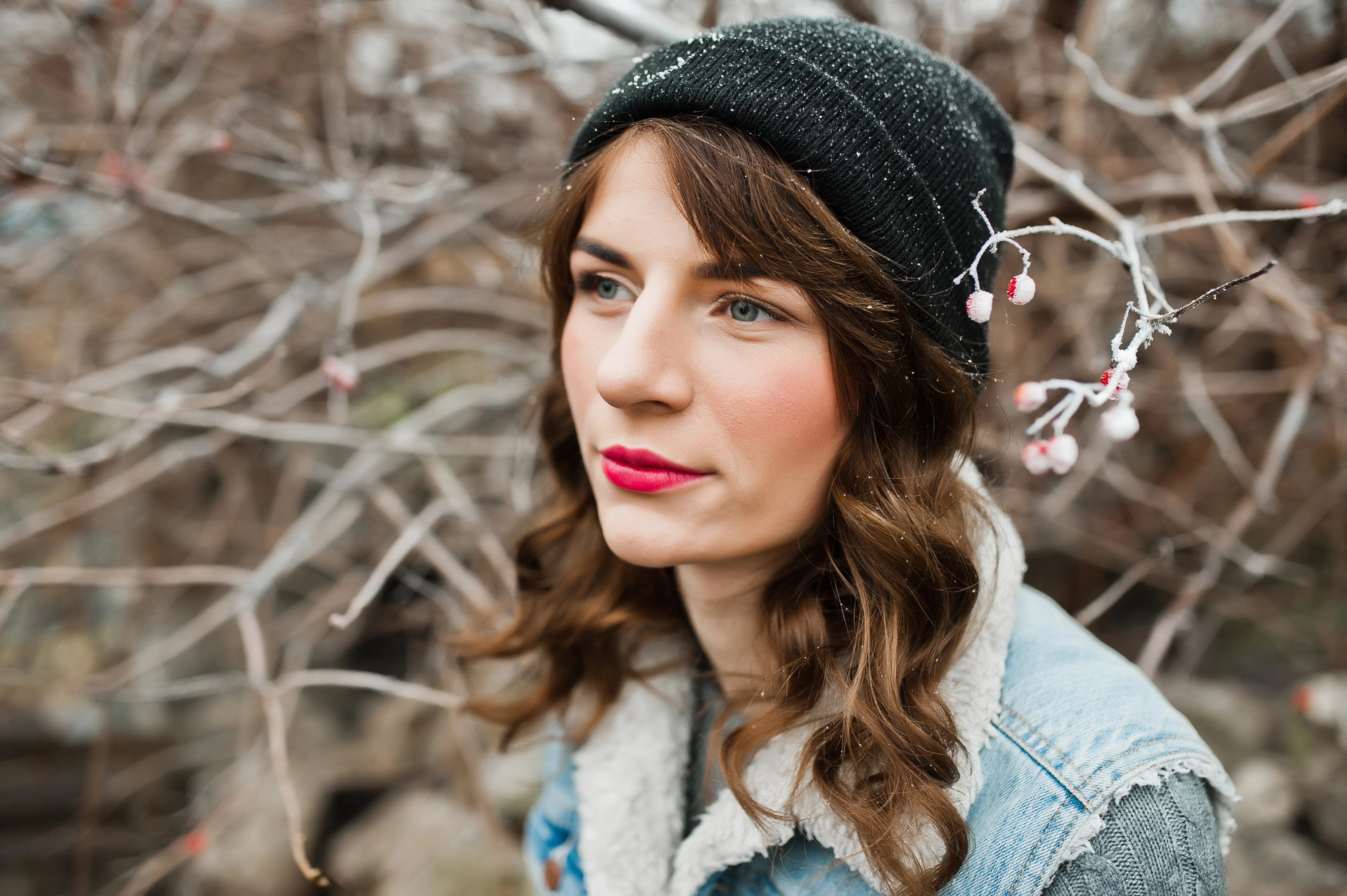 A person with long brown hair, red lipstick, and a black beanie stands outdoors in front of bare branches. They are wearing a denim jacket with a fleece collar and have a thoughtful expression. The branches have small buds on them.