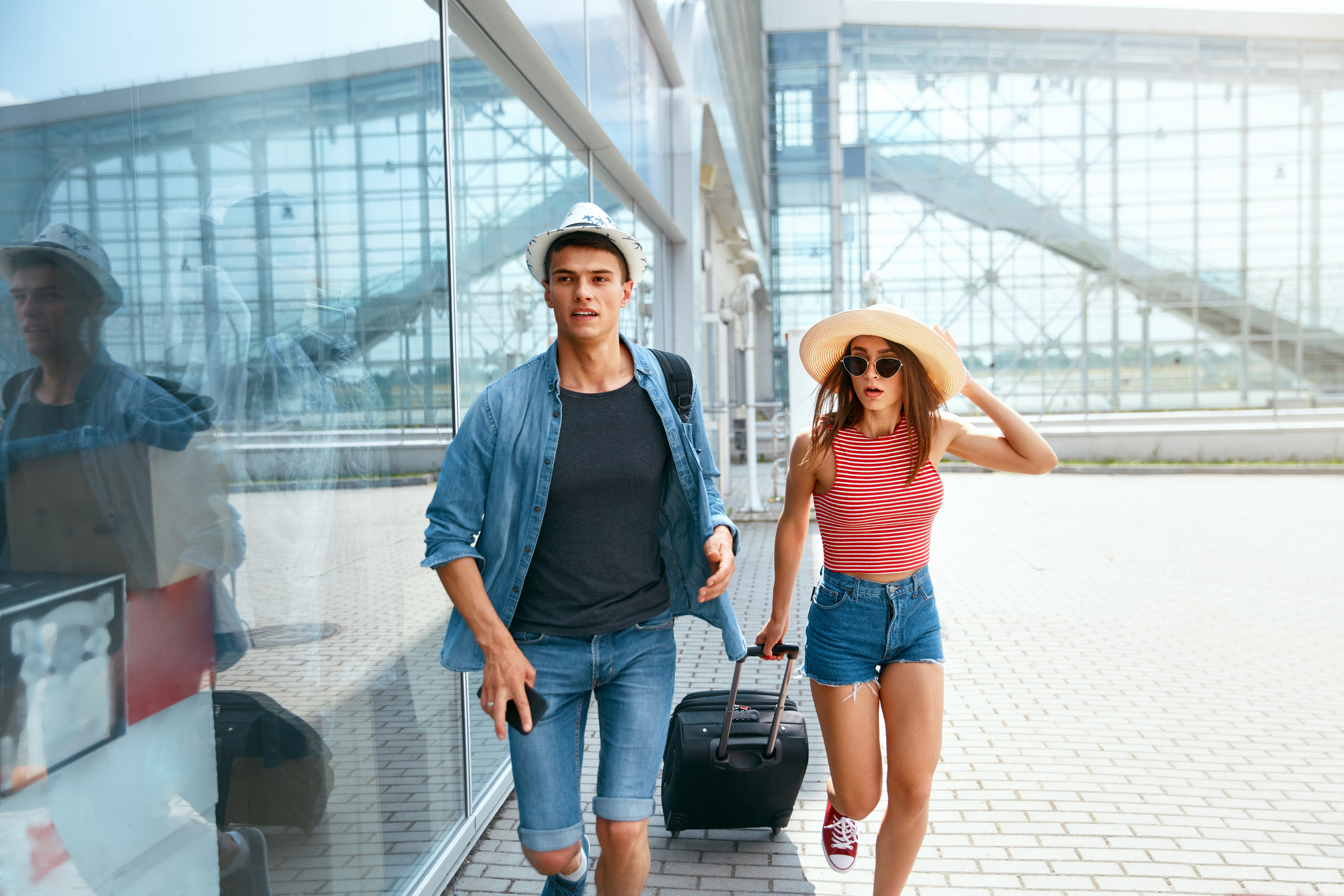 Two young adults, a man and a woman, walk outside an airport terminal. Both wear casual summer clothes and hats. The man carries a camera bag and the woman pulls a rolling suitcase. The terminal's glass wall reflects their images as they walk on the paved walkway.