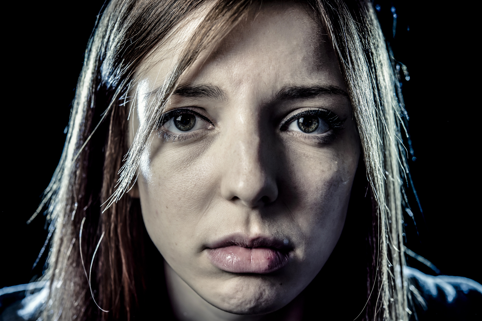 Close-up of a woman with light brown hair looking directly at the camera with a somber expression. Her eyes are slightly teary, and the lighting is dramatic, emphasizing the emotion in her face against a dark background.