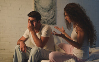 A man and a woman in loungewear are sitting on a bed against a white brick wall. The man is covering his face with one hand and appears distressed, while the woman, with curly hair, is gesturing emphatically with her hands, as if expressing strong emotions.