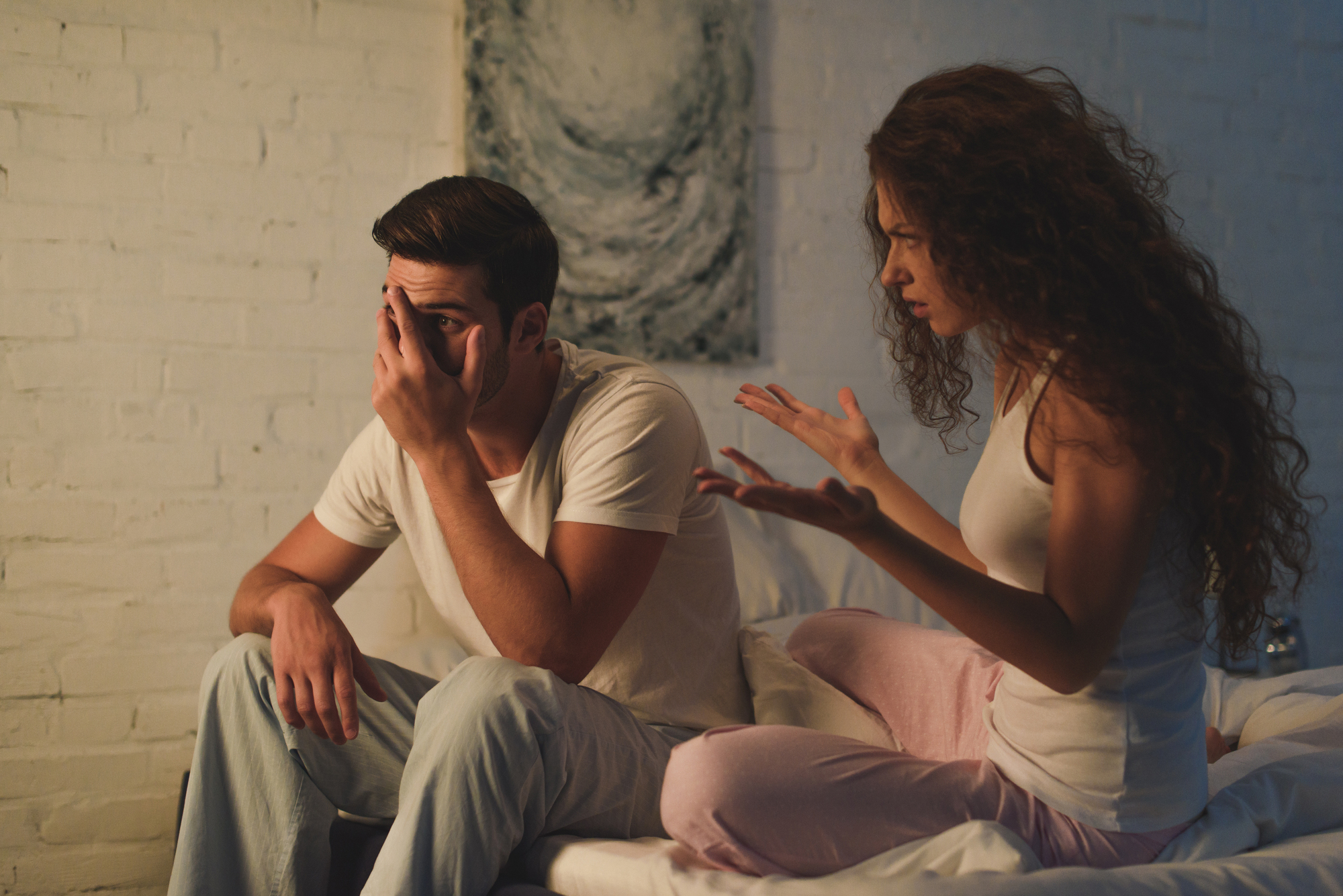 A man and a woman in loungewear are sitting on a bed against a white brick wall. The man is covering his face with one hand and appears distressed, while the woman, with curly hair, is gesturing emphatically with her hands, as if expressing strong emotions.