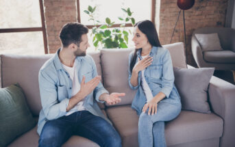 A man and a woman are sitting on a couch in a well-lit living room. Both are wearing denim outfits. They are engaged in a lively conversation, with the man gesturing and the woman placing a hand on her chest. There's a plant and a large window in the background.