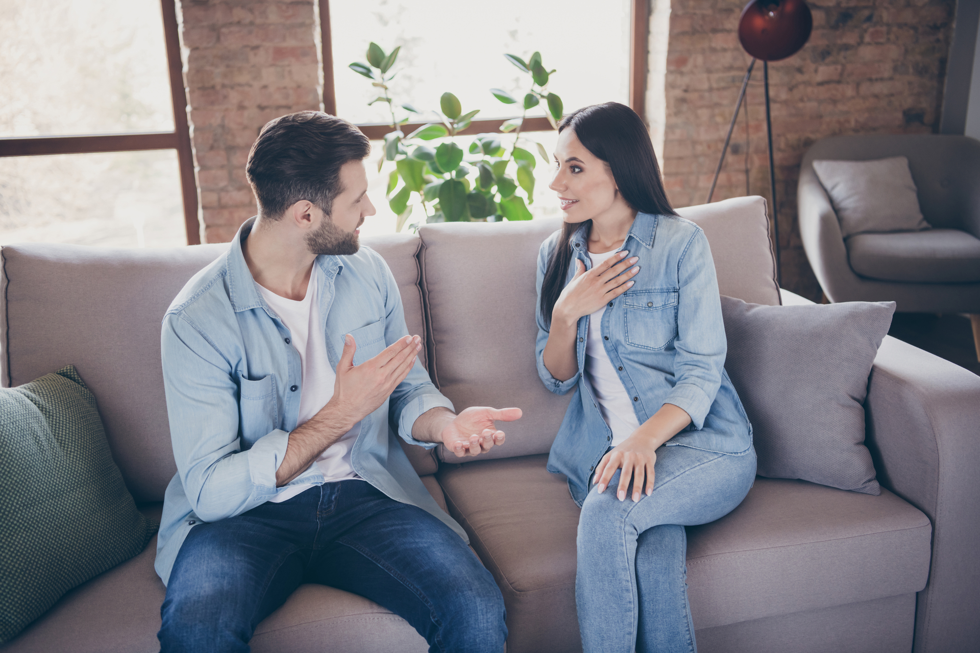 A man and a woman are sitting on a couch in a well-lit living room. Both are wearing denim outfits. They are engaged in a lively conversation, with the man gesturing and the woman placing a hand on her chest. There's a plant and a large window in the background.