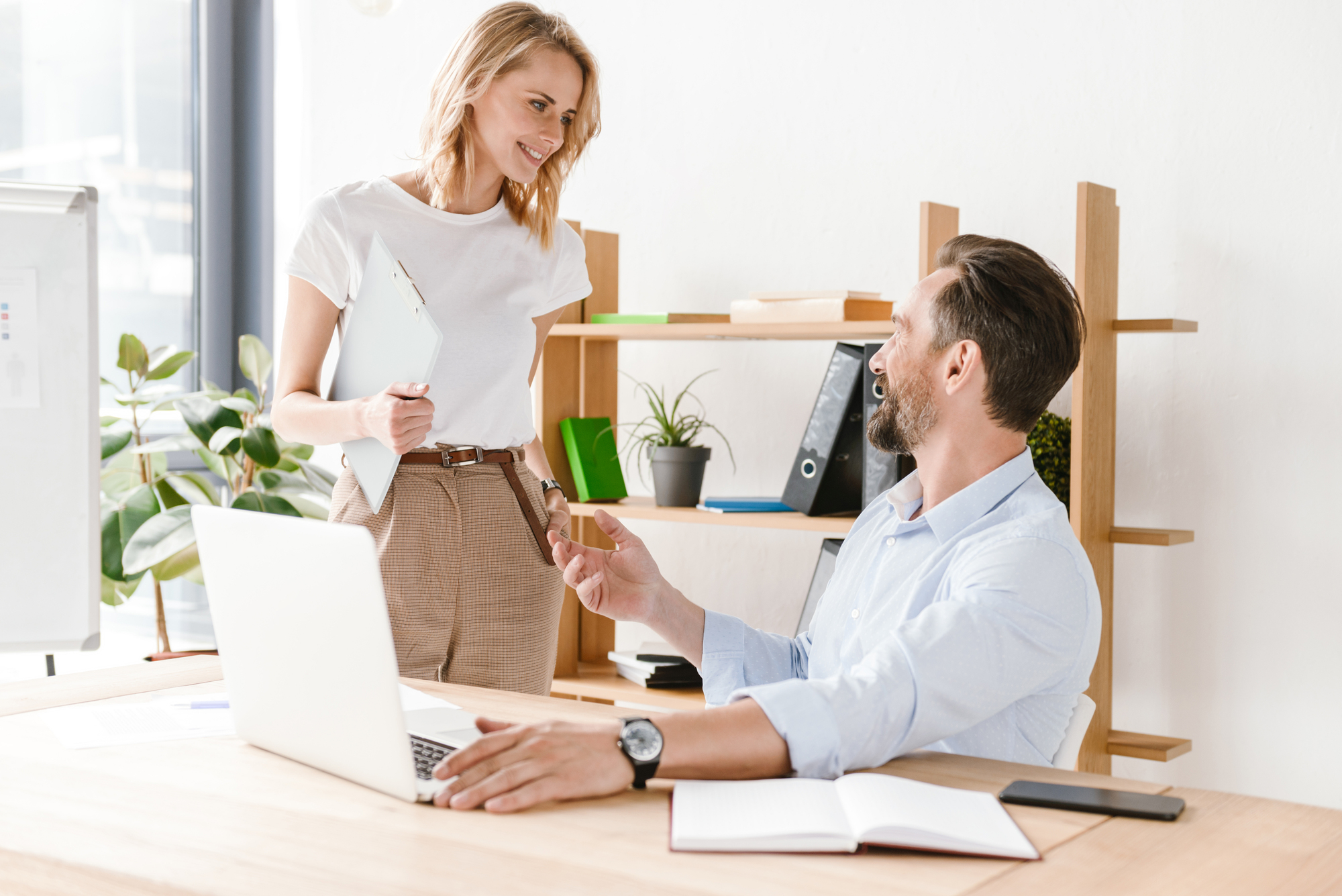 A woman holding a clipboard smiles and engages in conversation with a seated man who is holding a smartphone. They are in a modern office setting, with a laptop, notebook, and bookshelf filled with various items in the background.