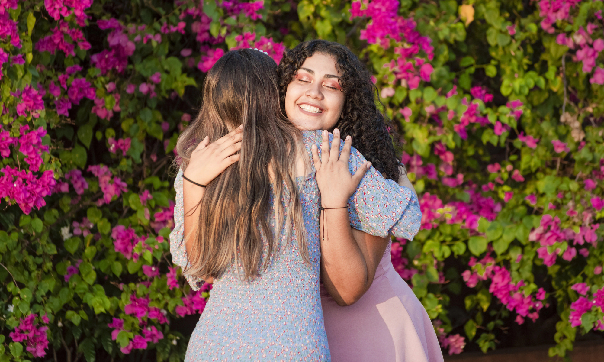 Two women embrace joyfully in front of a vibrant background of pink bougainvillea flowers and green leaves. One has curly hair and wears a light blue dress; the other has long hair and wears a blue dress with a floral pattern. Both are smiling and appear happy.