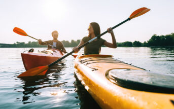 Two people are kayaking on a calm body of water. The person in the foreground is in a yellow kayak, and the one in the background is in an orange kayak. Both are holding paddles and wearing sunglasses, enjoying a sunny day with trees visible in the distance.