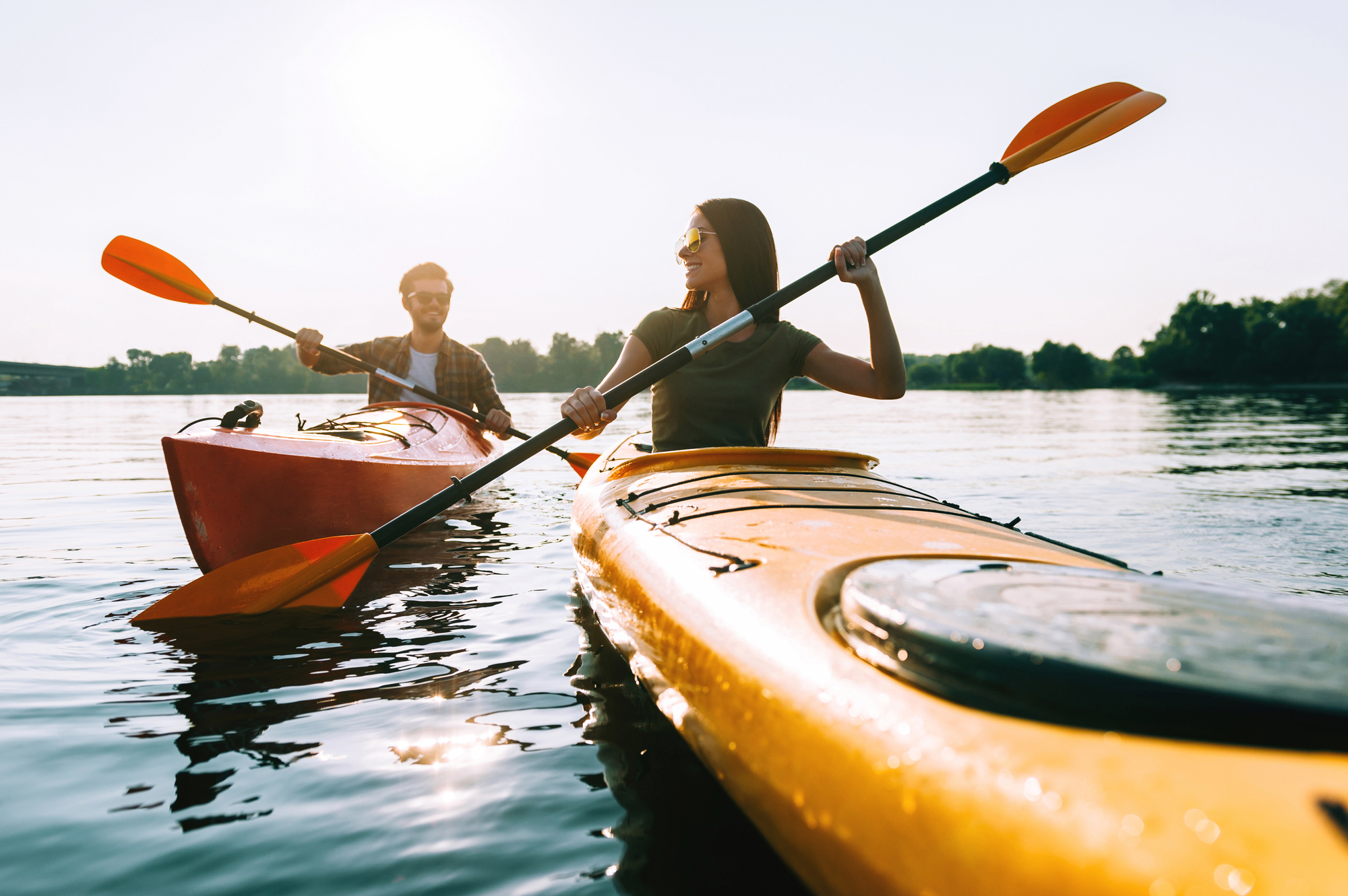 Two people are kayaking on a calm body of water. The person in the foreground is in a yellow kayak, and the one in the background is in an orange kayak. Both are holding paddles and wearing sunglasses, enjoying a sunny day with trees visible in the distance.