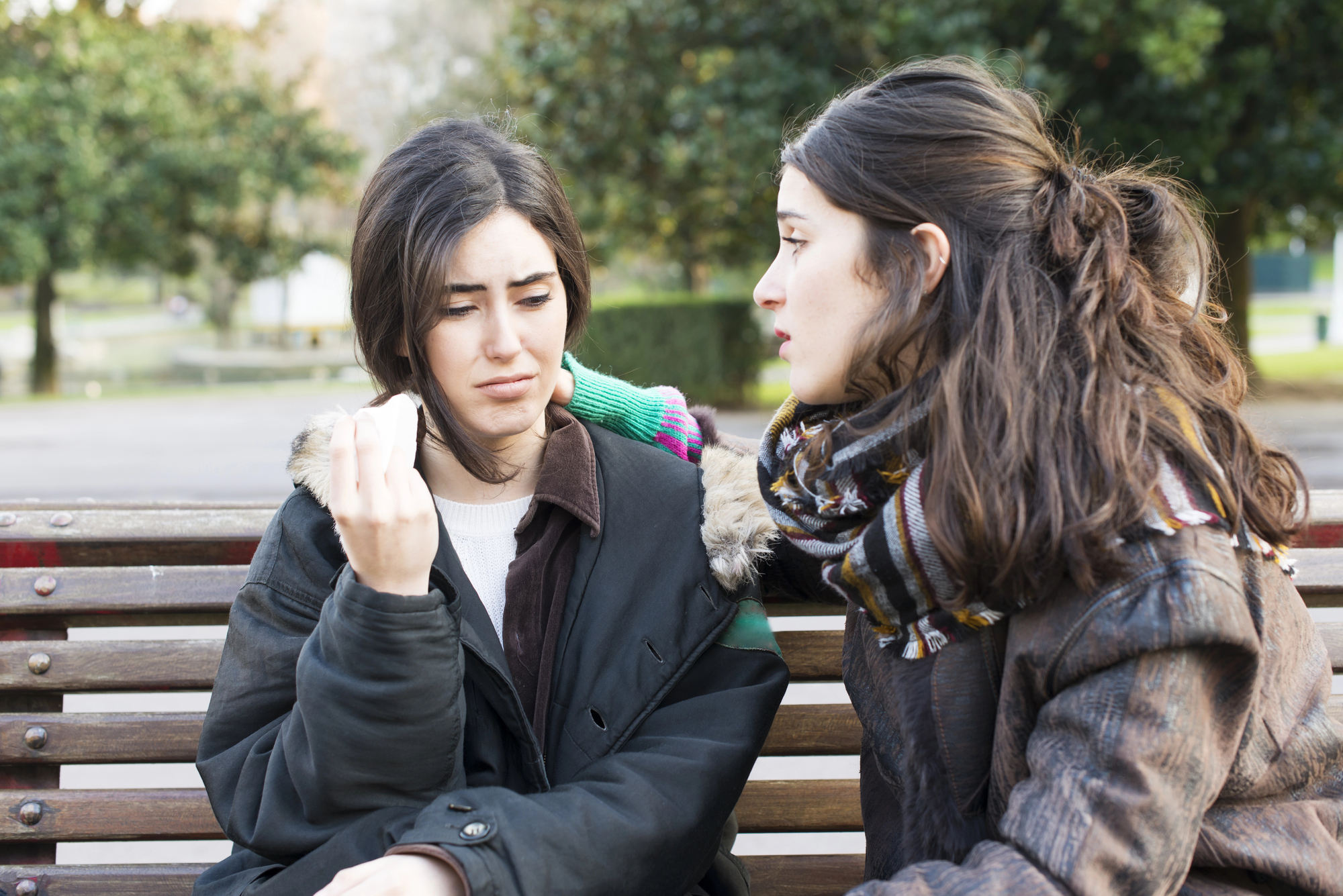 Two women sit on a park bench. The woman on the left appears upset, holding a tissue to wipe her tears, while the woman on the right, wearing a colorful scarf, offers comfort by placing a hand on her shoulder. Trees and a path are visible in the background.