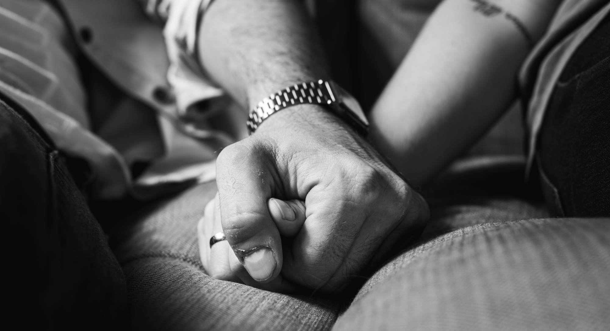 Black-and-white image of two people holding hands. One person has a wristwatch and a wedding ring on their left hand, while the other person has a tattoo on their forearm. The image focuses on their interlocking hands, conveying a sense of connection and support.