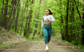 A woman in athletic wear, including a white crop top and green leggings, is running on a dirt path through a forest. The trees around her are lush and green, suggesting it is spring or summer. She appears to be focused and determined.