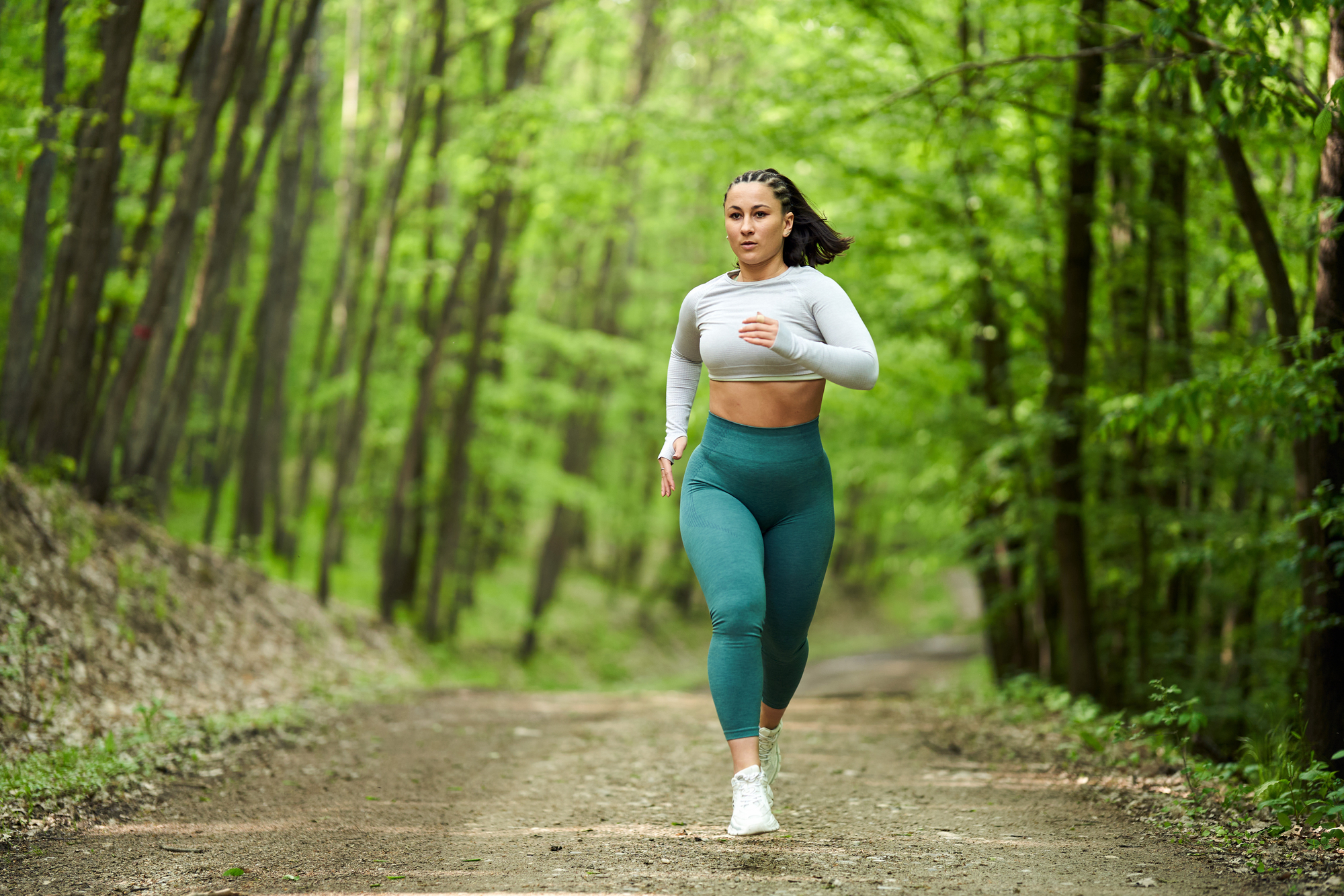 A woman in athletic wear, including a white crop top and green leggings, is running on a dirt path through a forest. The trees around her are lush and green, suggesting it is spring or summer. She appears to be focused and determined.