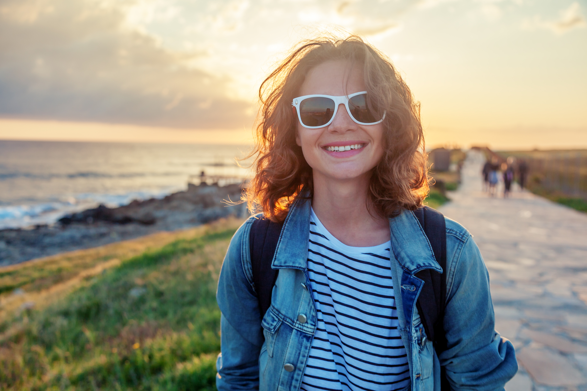 A person with curly hair, wearing white sunglasses, a denim jacket, and a striped shirt smiles at the camera. They are standing on a pathway by the sea at sunset, with a rocky shoreline and silhouettes of people in the background.