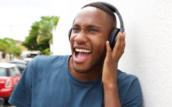 A person wearing headphones leans against a white wall outdoors, smiling widely with their mouth open. They are wearing a blue t-shirt and appear to be enjoying the music. A blurred outdoor scene with trees and a red car is in the background.