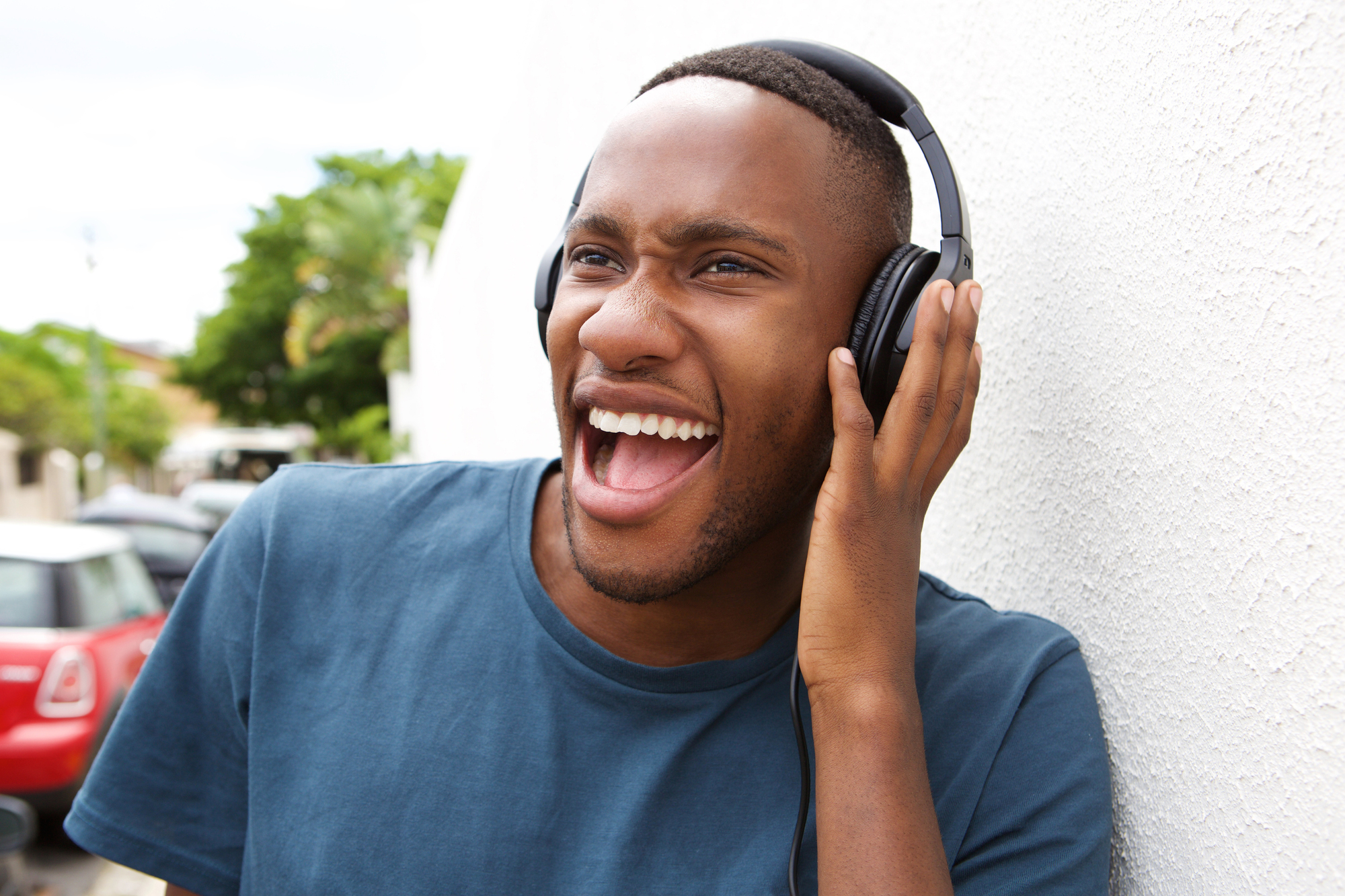 A person wearing headphones leans against a white wall outdoors, smiling widely with their mouth open. They are wearing a blue t-shirt and appear to be enjoying the music. A blurred outdoor scene with trees and a red car is in the background.