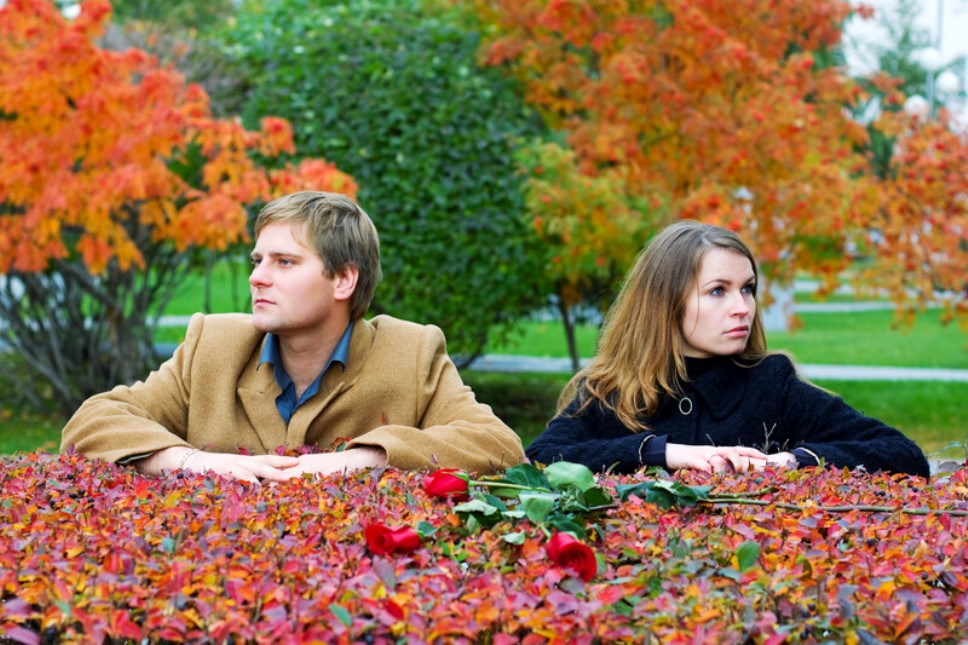 Two individuals, a man and a woman, sit opposite each other at a table adorned with red roses and autumn leaves. Both appear contemplative, gazing in opposite directions. The background features vibrant fall foliage, adding a warm, seasonal ambiance.