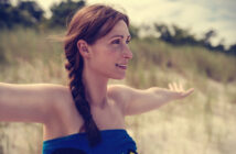 A woman with a braid stands outdoors with her arms outstretched, smiling and looking to the side. She is wrapped in a blue towel and has a background of tall grass and trees. The weather appears to be sunny.